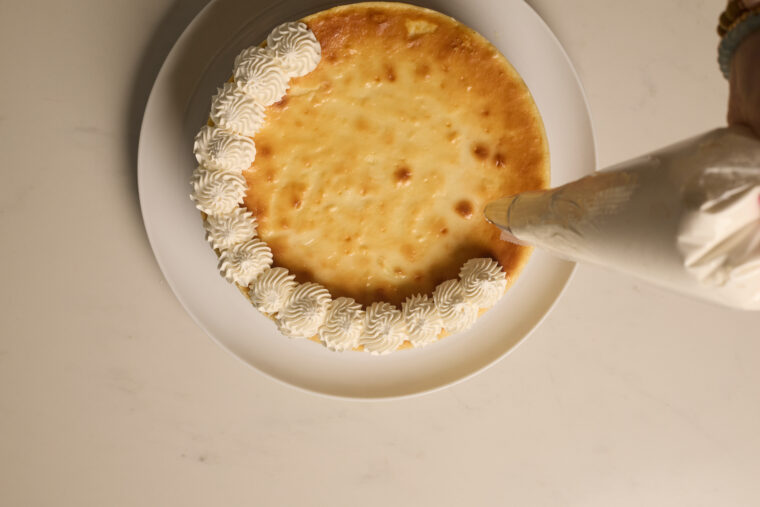 Someone&rsquo;s piping fluffy whipped cream onto the edges of a strawberry cheesecake sitting on a white plate.