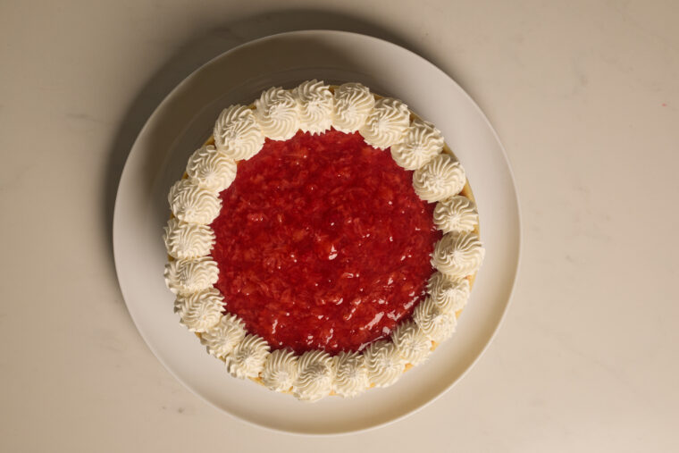 Strawberry cheesecake with a red fruity glaze and whipped cream on the edges, sitting on a white plate against a pale background.