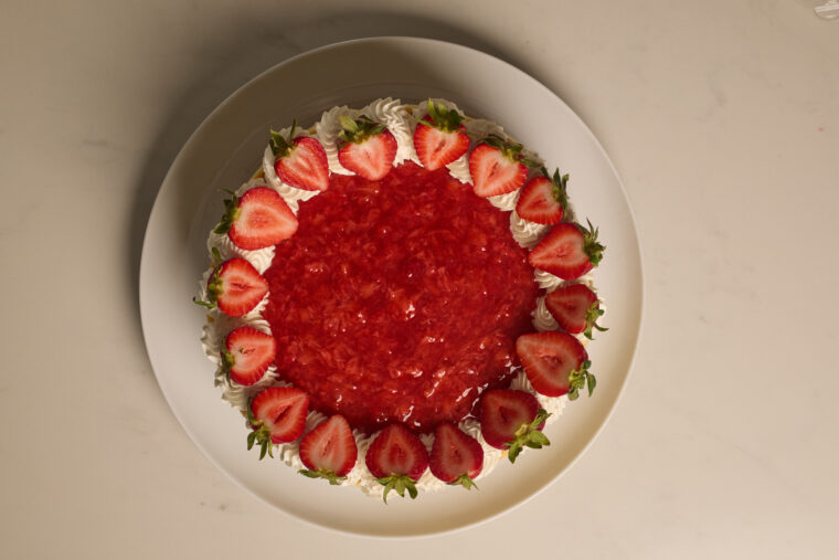 A creamy, round strawberry cheesecake topped with whipped cream sits on a white plate, seen from above on a light background.