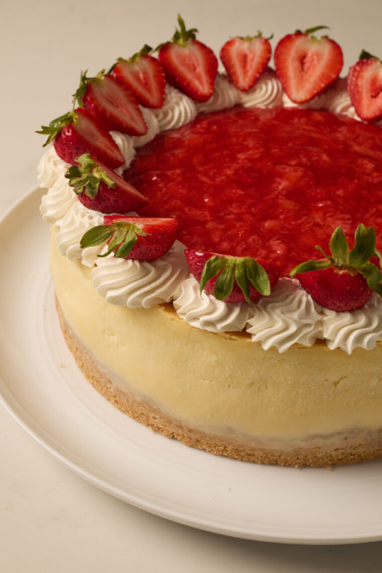 A rich strawberry cheesecake sits on a plate, loaded with whipped cream, white chocolate, strawberry sauce, and fresh berries.
