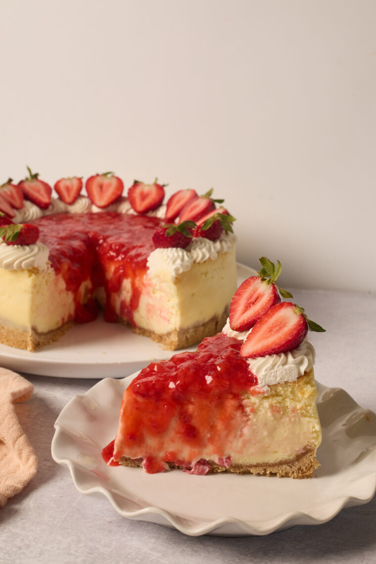 A yummy slice of strawberry cheesecake with sauce and whipped cream on a plate, the rest of the cake behind it.