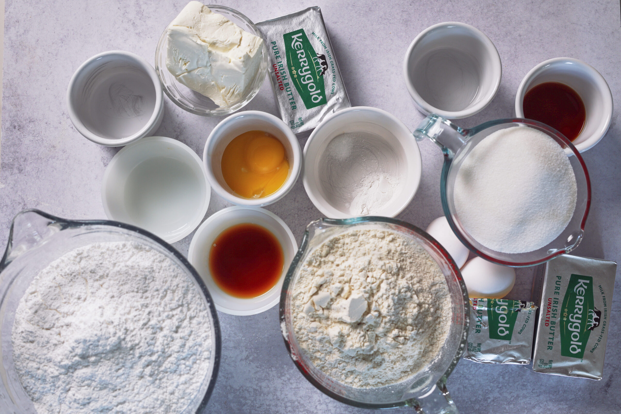 A bunch of baking staples&mdash;flour, sugar, eggs, butter, cream cheese, vanilla, and little bowls&mdash;are spread out on a pale counter.