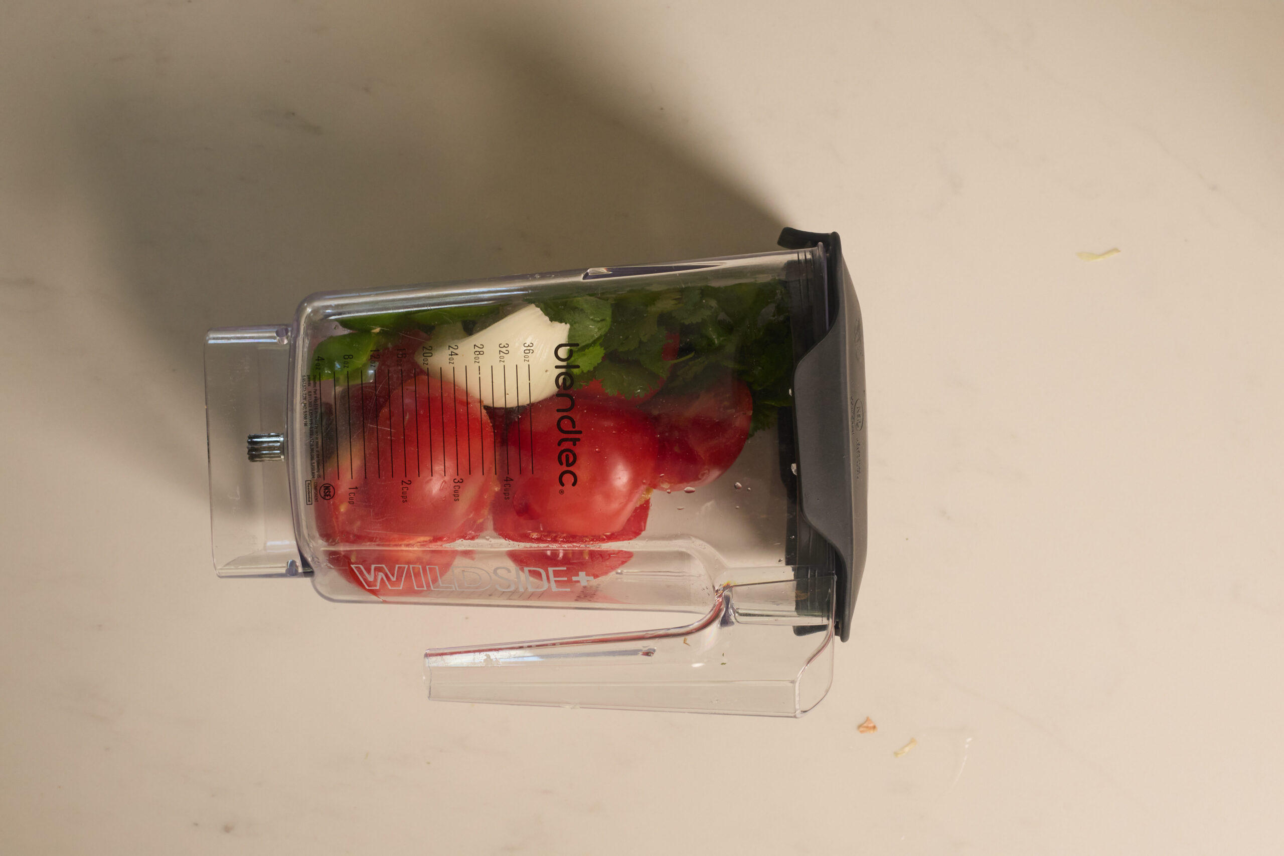 A blender cup packed with tomatoes, cilantro, and garlic sits on a pale counter, all set for homemade salsa.