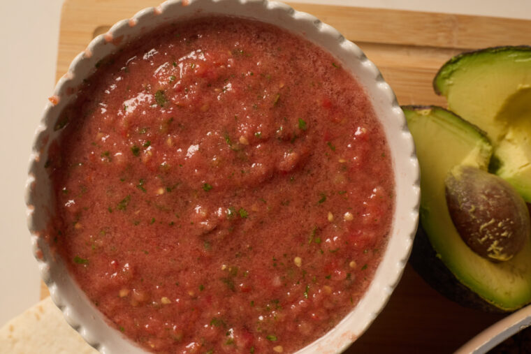 A bowl of bright red salsa with herbs is on a wooden board right next to a freshly cut avocado half.