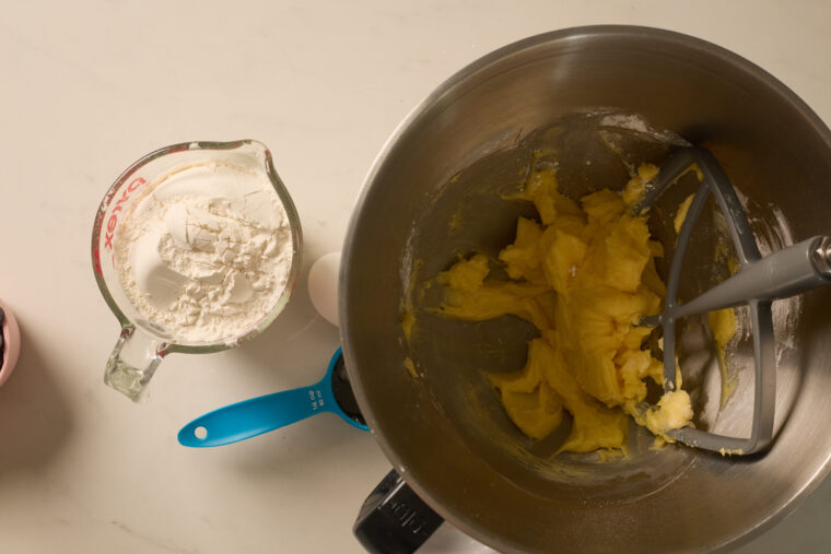 Mixing bowl with creamed butter and sugar, flour, a blue spoon, and an egg on the counter&mdash;all set for shortbread cookies!.