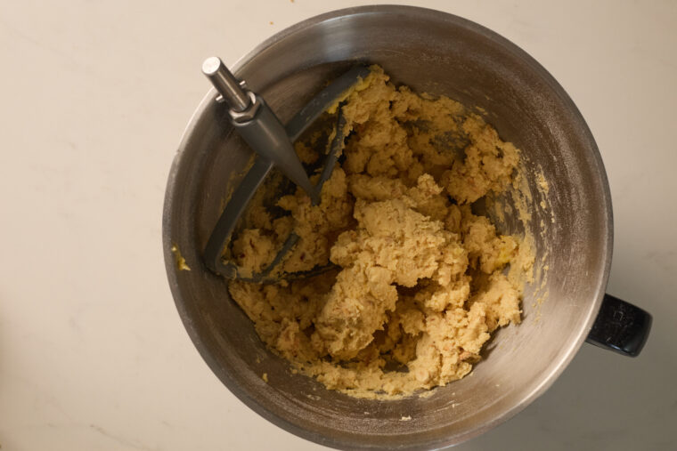 Looking down at a bowl of thick shortbread dough with a paddle attachment, sitting on a light countertop.