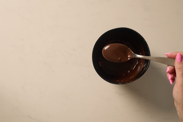 Someone with pink nails is about to drizzle melted chocolate from a spoon over shortbread cookies on a bright counter.