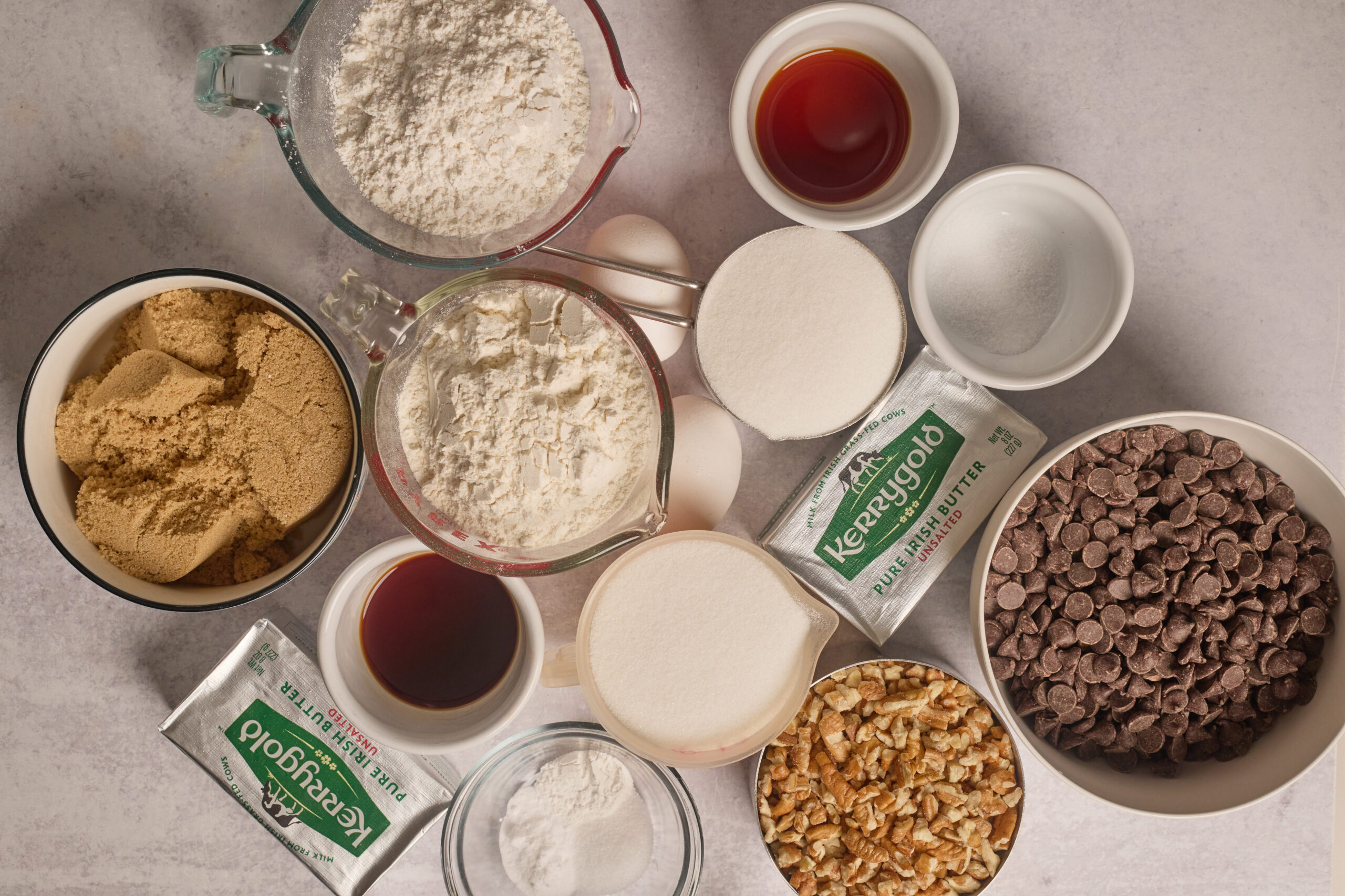 A bunch of baking stuff on the counter—flour, eggs, butter, sugars, vanilla, baking soda, salt, choc chips, and walnuts.