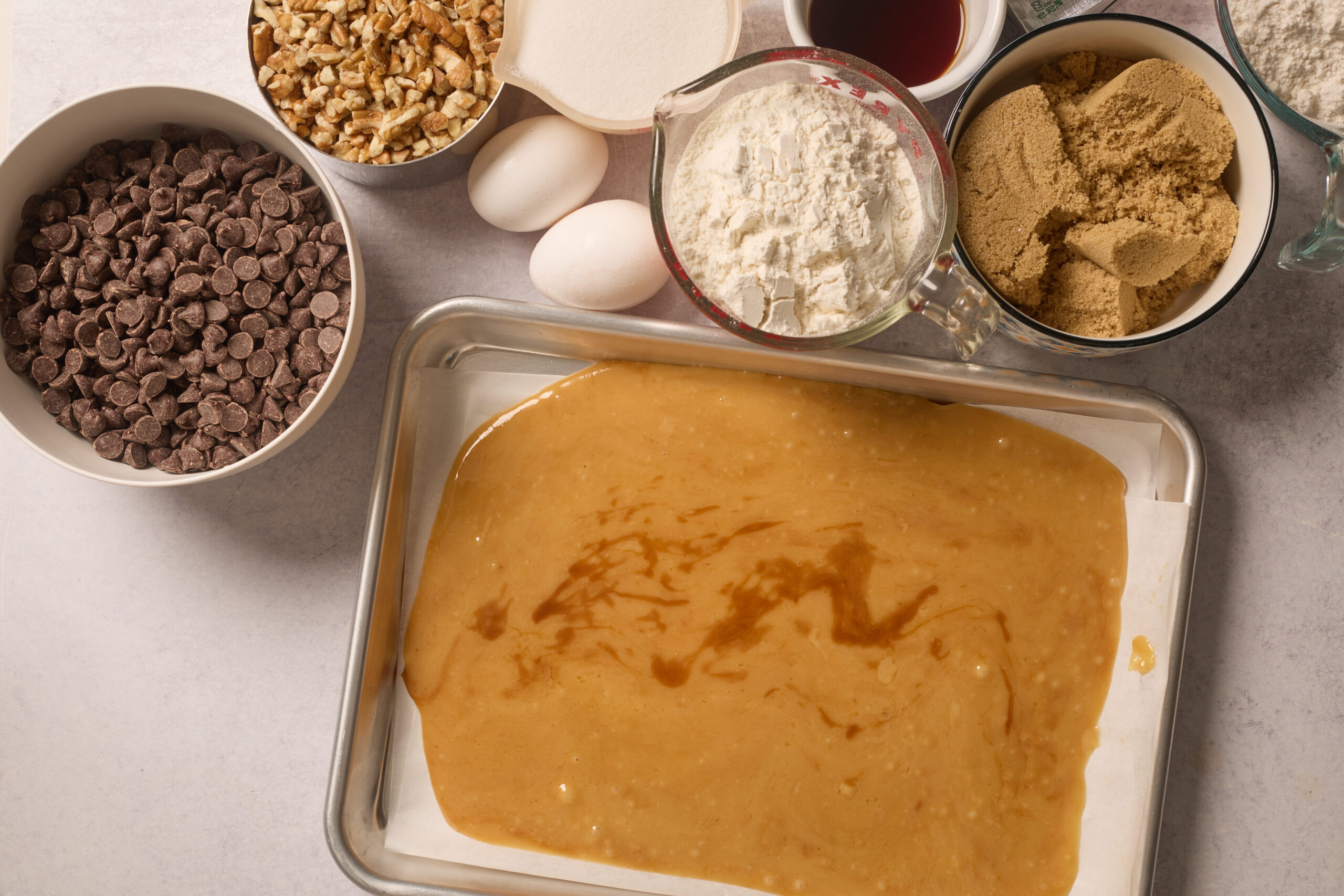 A baking tray filled with batter sits on a counter, surrounded by bowls of chocolate chips, nuts, eggs, flour, sugar, and vanilla.