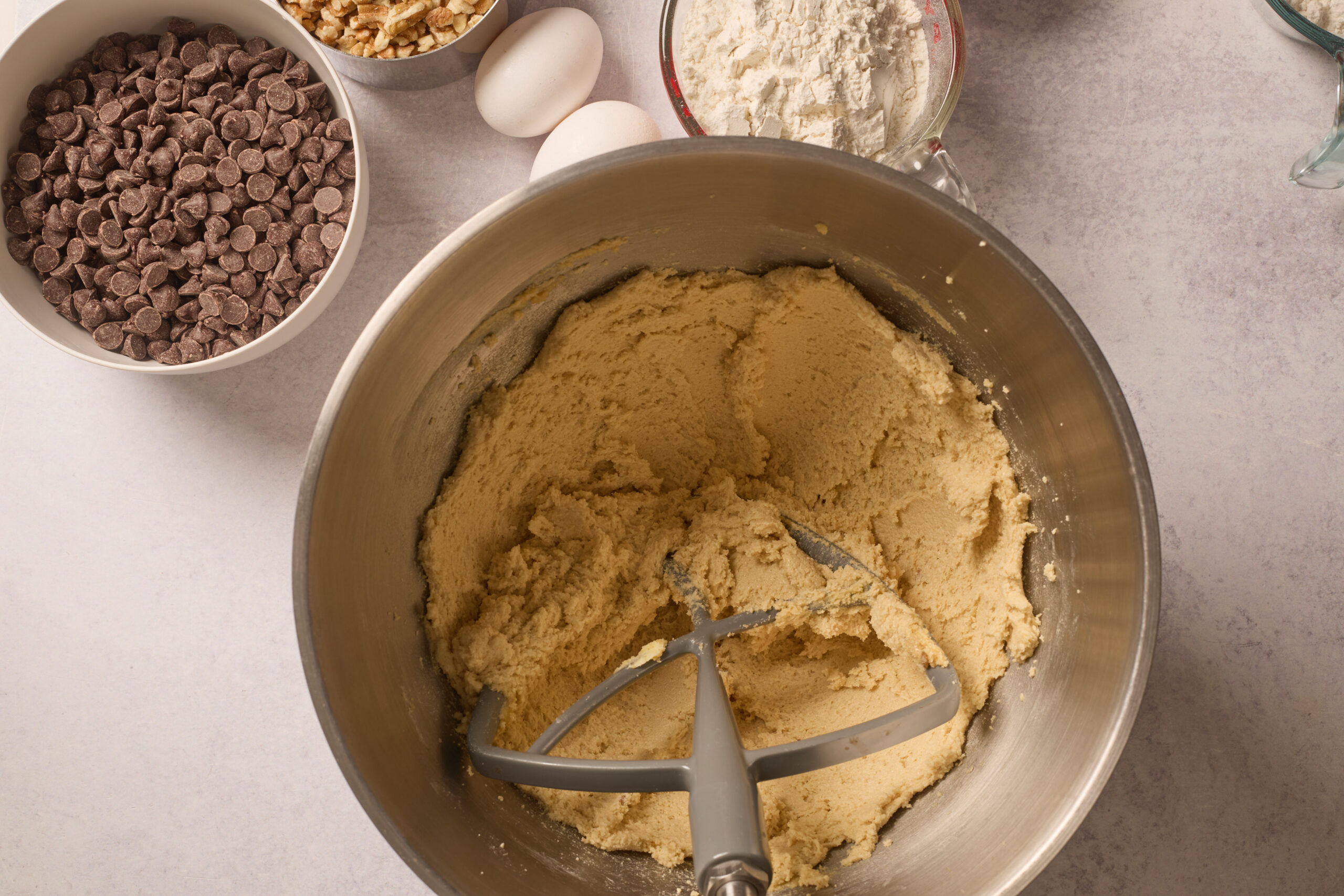 A metal bowl of cookie dough with a mixer paddle, plus chocolate chips, walnuts, eggs, and flour scattered on a light counter.