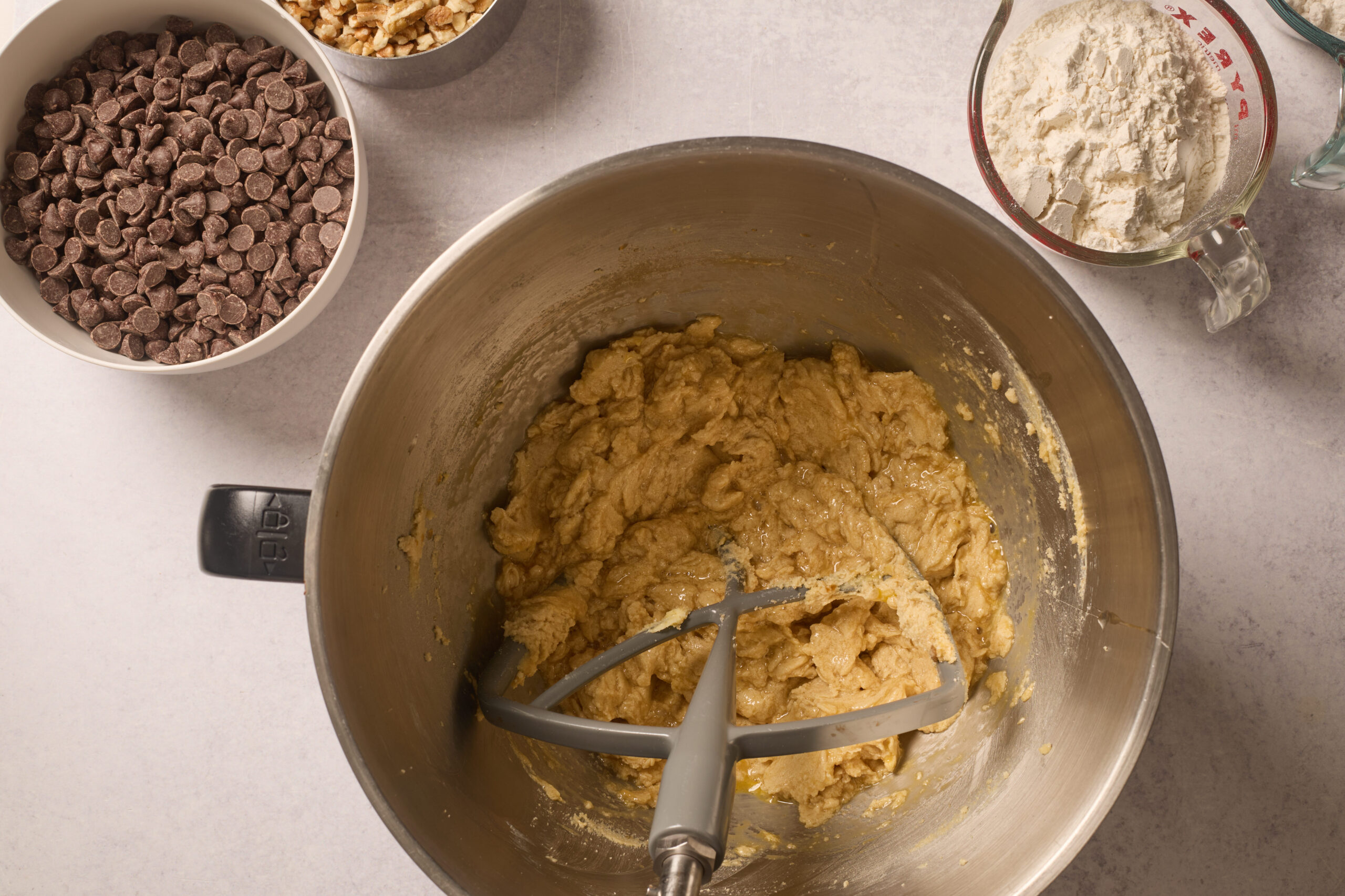 A mixing bowl with cookie dough and mixer sits on the counter, near chocolate chips, nuts, and a flour-filled measuring cup.