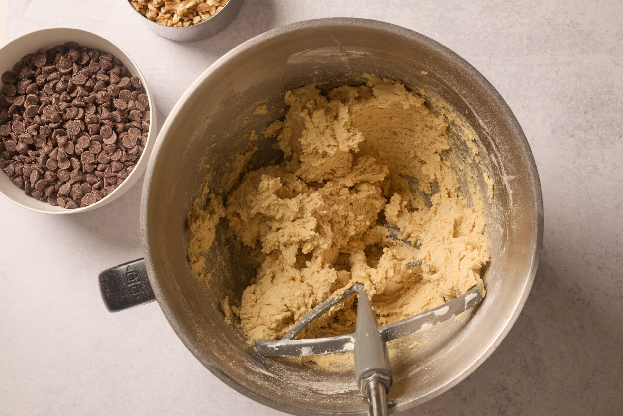 Cookie dough in a bowl with a metal mixer sits nearby chocolate chips and nuts on a light-colored surface.