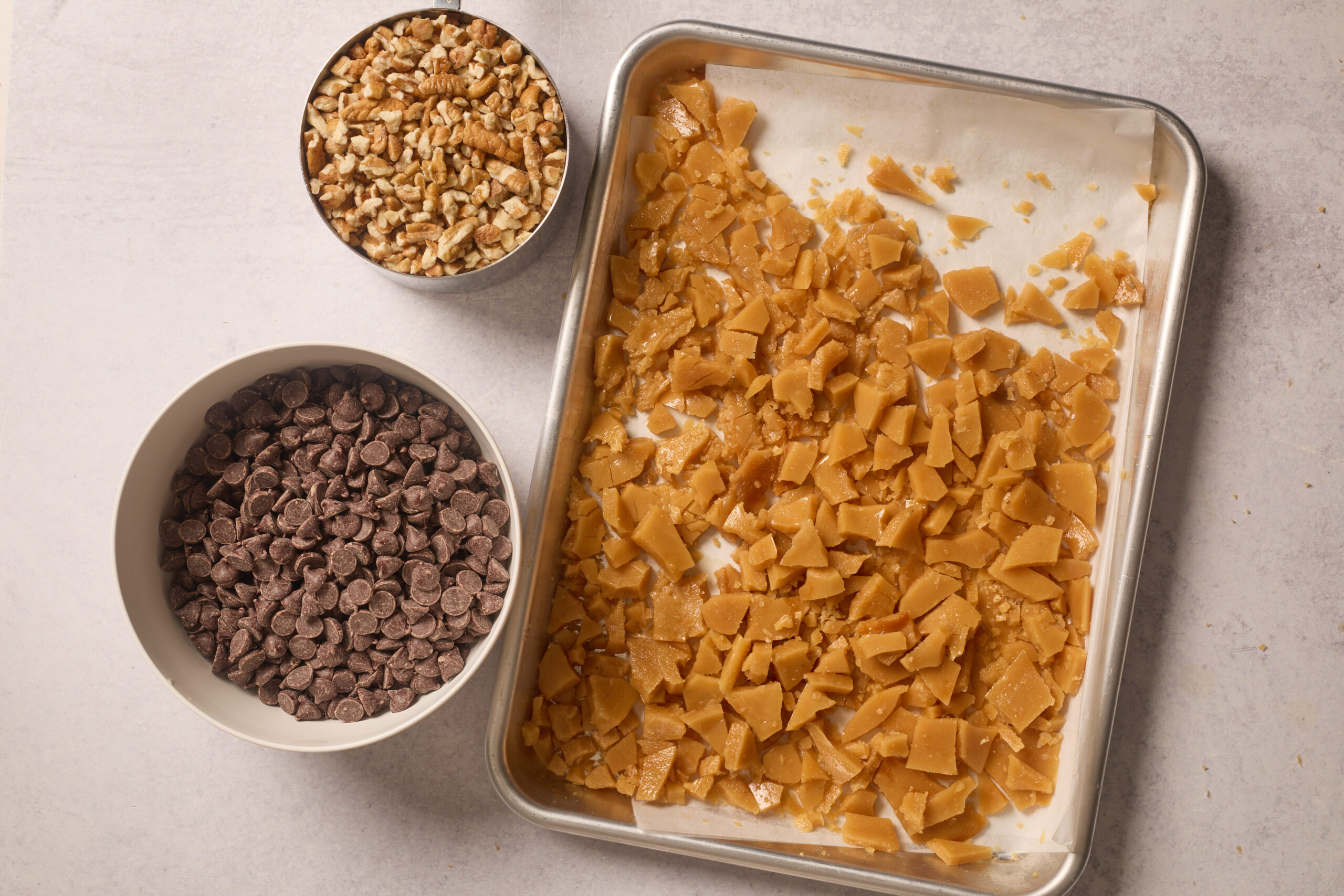 Broken toffee on a baking tray sits beside bowls of chocolate chips and chopped nuts, all spread out on a light countertop.