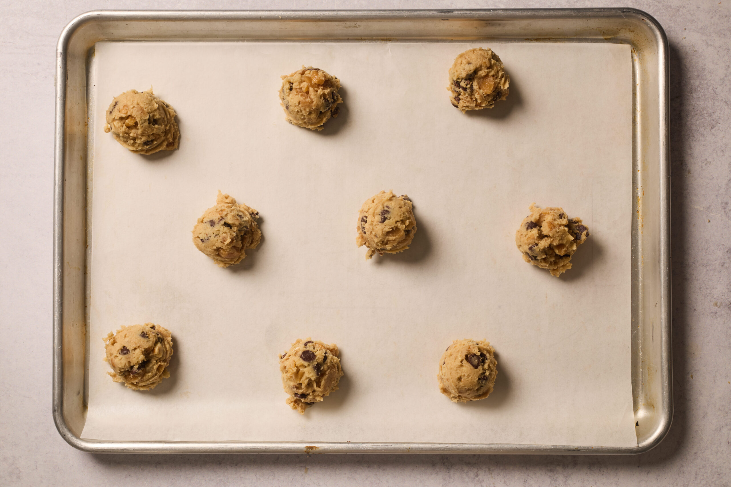 Nine balls of chocolate chip cookie dough are spread out on a baking sheet lined with parchment paper.
