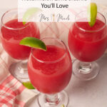 Three glasses of bright watermelon-lime slushie with lime wedges sit on a light table with sliced limes and a pink checkered cloth.
