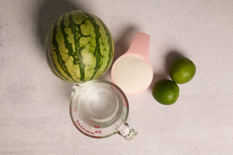 A whole watermelon, two limes, water in a glass cup, and sugar in a pink cup sit on a light surface&mdash;slushi time!.