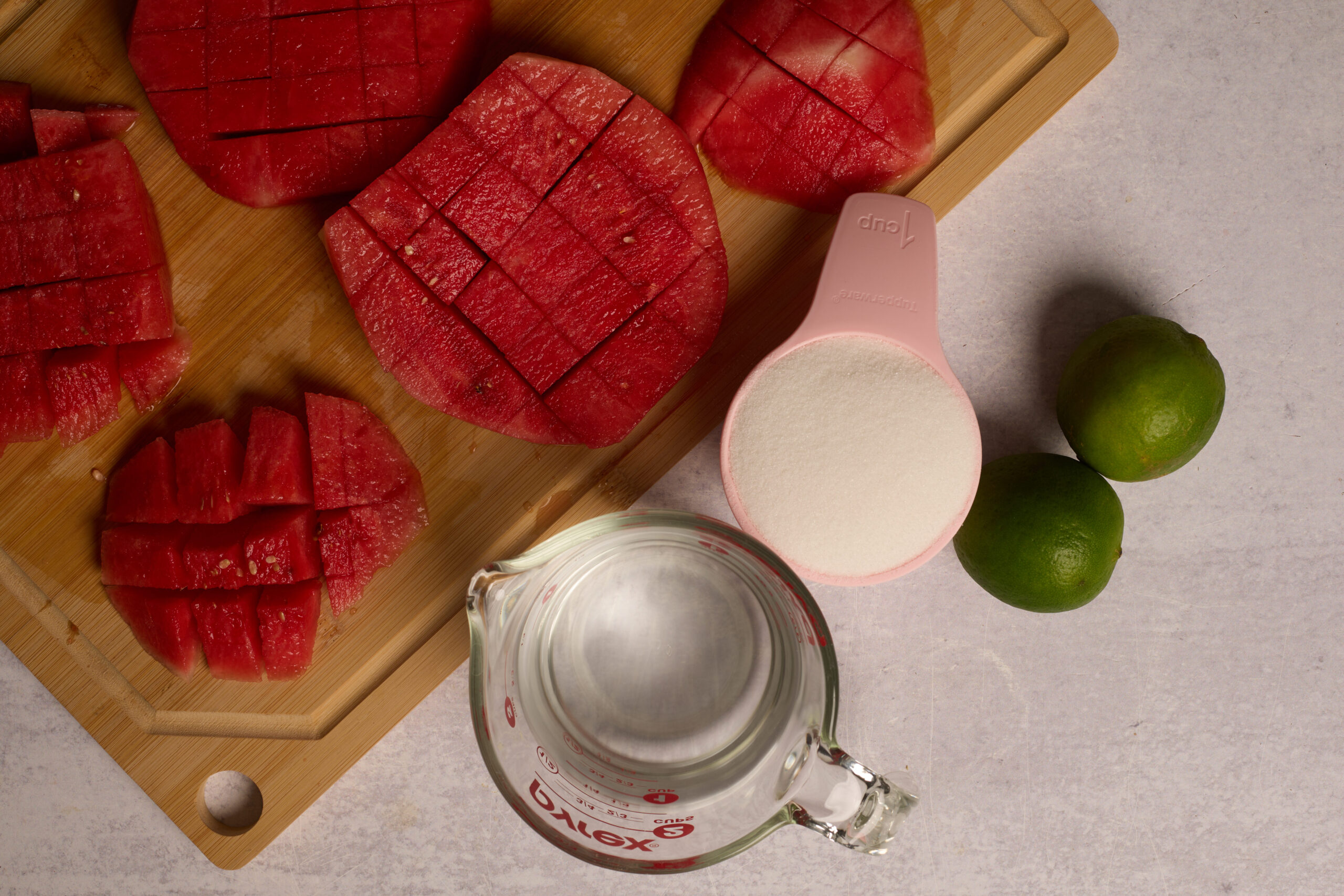 Chunks of watermelon on a board, sugar in a cup, water in a measuring glass, and two limes hanging out on a light counter.