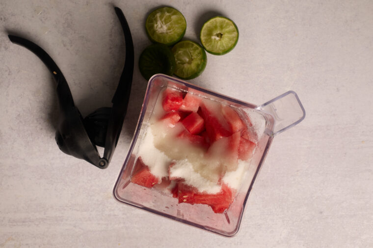 A blender with watermelon, sugar, and ice sits near a citrus squeezer and lime slices, ready for a tasty slushie.