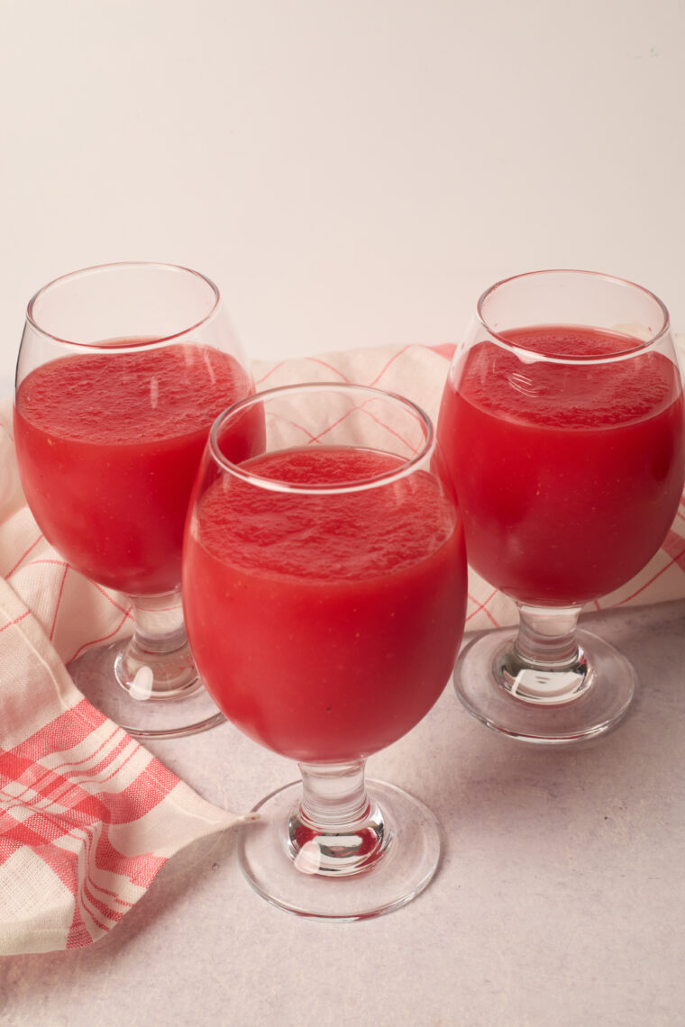 Three fancy glasses of rich red watermelon juice sit on a light table by a pink-and-white checkered cloth.
