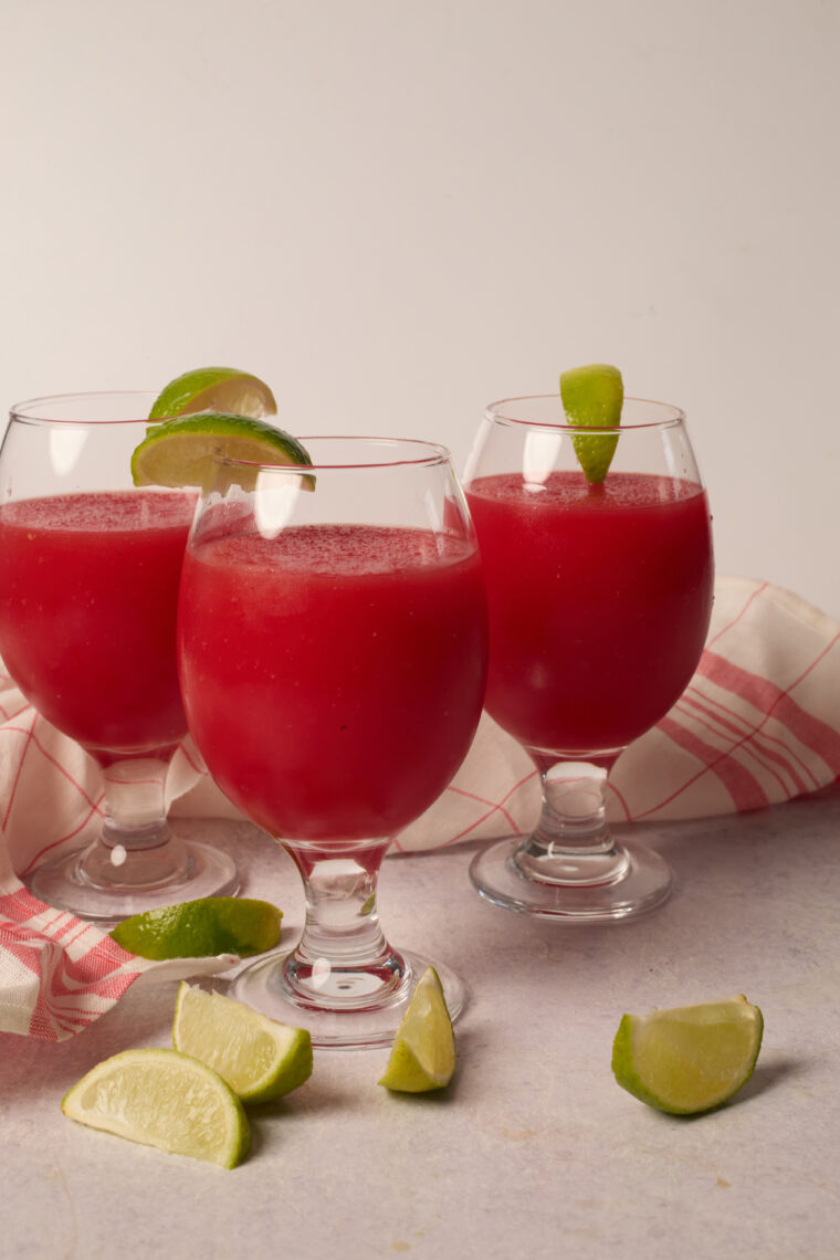 Three glasses of watermelon slush, each with a lime wedge, on a white table with a red-striped cloth and some lime slices.