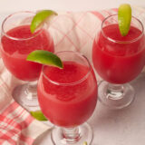 Three watermelon slushies with lime wedges are on a light table by a pink checkered cloth, plus some extra limes nearby.