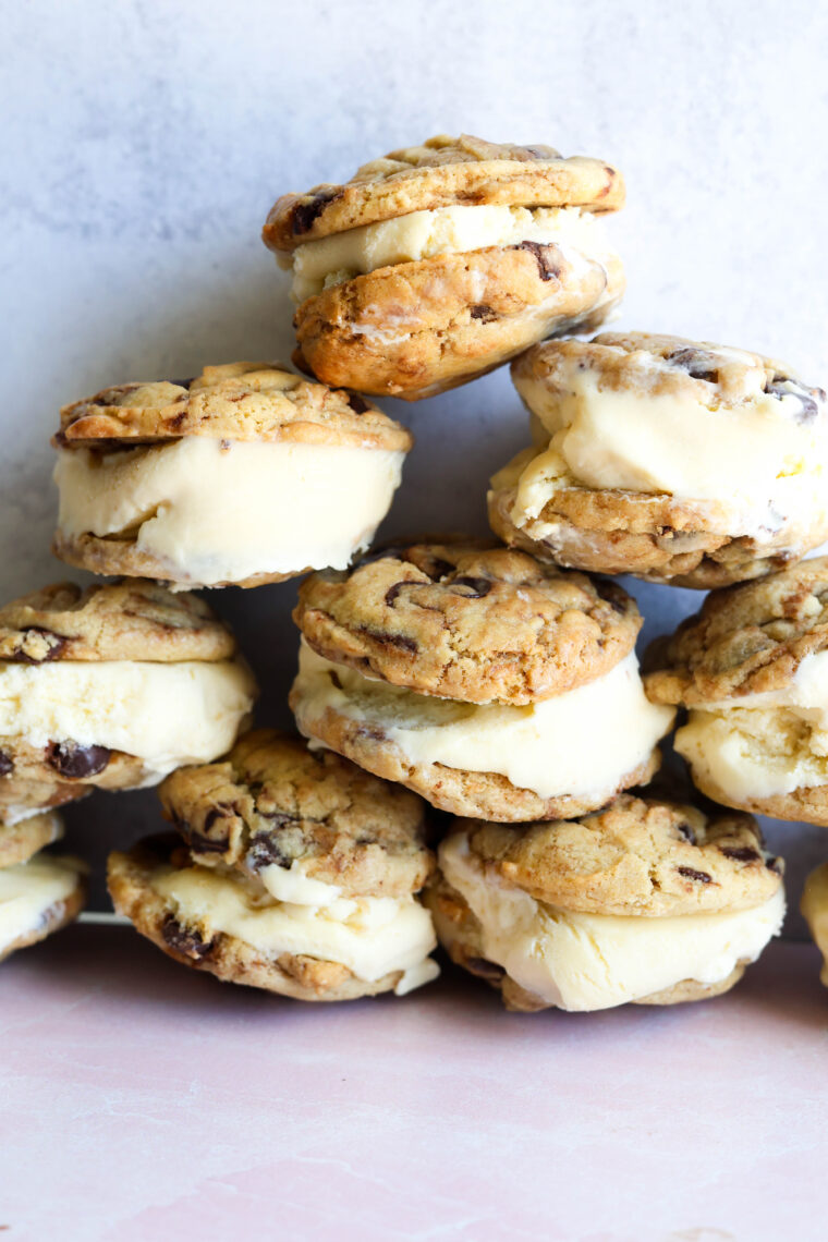 A pile of vanilla chocolate chip cookie ice cream sandwiches sits on a light pink surface with a plain white backdrop.