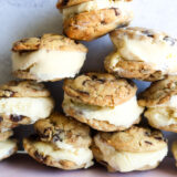 A pile of vanilla chocolate chip cookie ice cream sandwiches sits on a light pink surface with a plain white backdrop.