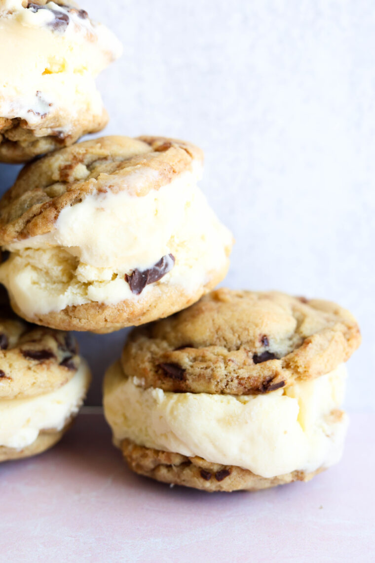 Three chocolate chip cookie ice cream sandwiches are stacked up on a light surface. Each one has creamy vanilla ice cream tucked between two cookies—a fun twist on the classic treat!.