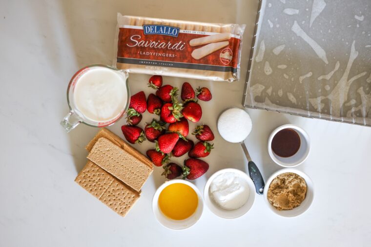 All the fixings for No-Bake Strawberry Shortcake Icebox Bars are spread out on the counter: fresh strawberries, ladyfingers, graham crackers, milk, honey, both white and brown sugar, vanilla extract, plus a metal pan lined with parchment paper ready to go.