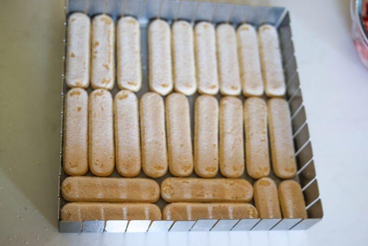 A square pan sits on a light countertop, filled with rows of dry ladyfinger cookies. It’s all set up and ready for making some easy, no-bake Strawberry Shortcake Icebox Bars.