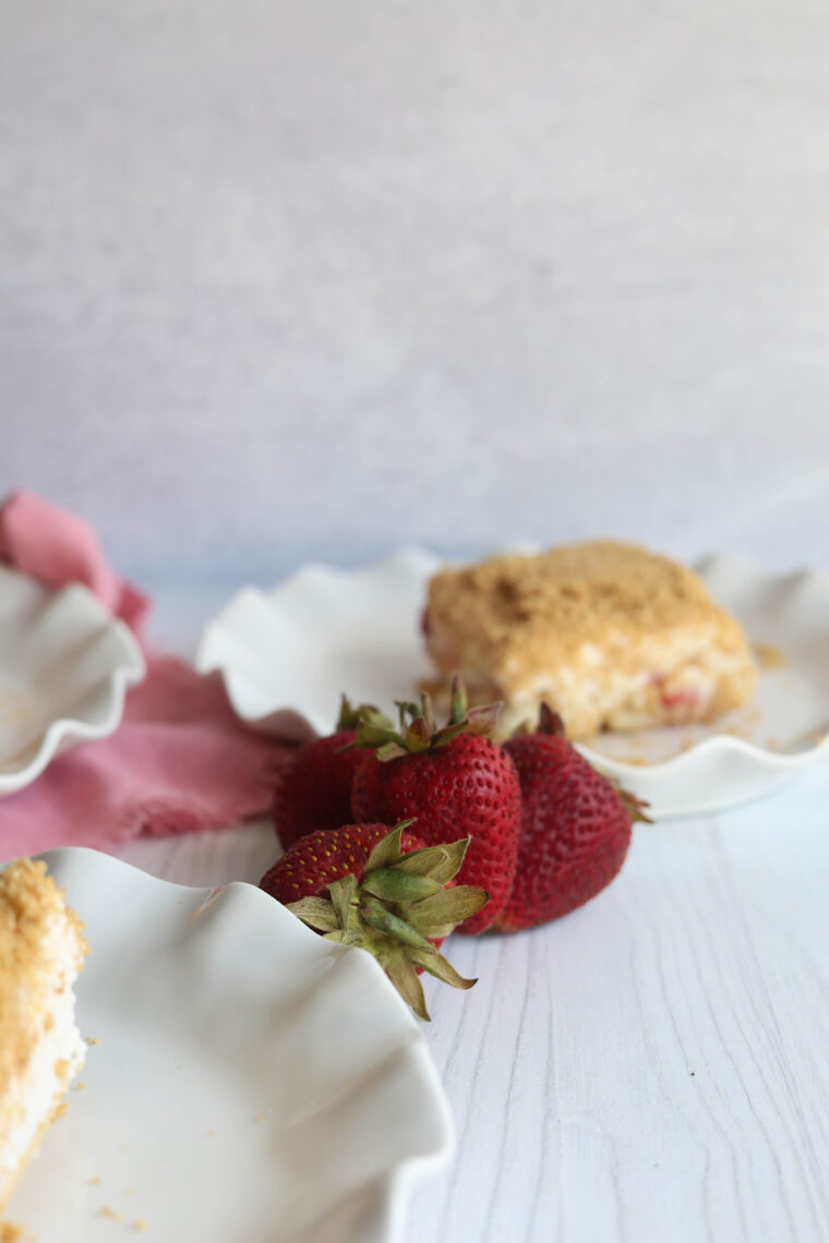 Three strawberries sit on a white surface next to plates piled with crumbly-topped strawberry shortcake. There’s a pink cloth hanging out in the background.