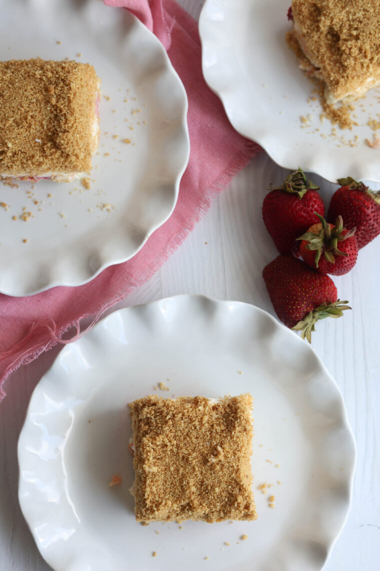 Three plates each have a piece of No-Bake Strawberry Shortcake Icebox Bar with some crumbly topping, sitting on a white table. A pink cloth is nearby and there are three fresh strawberries too.