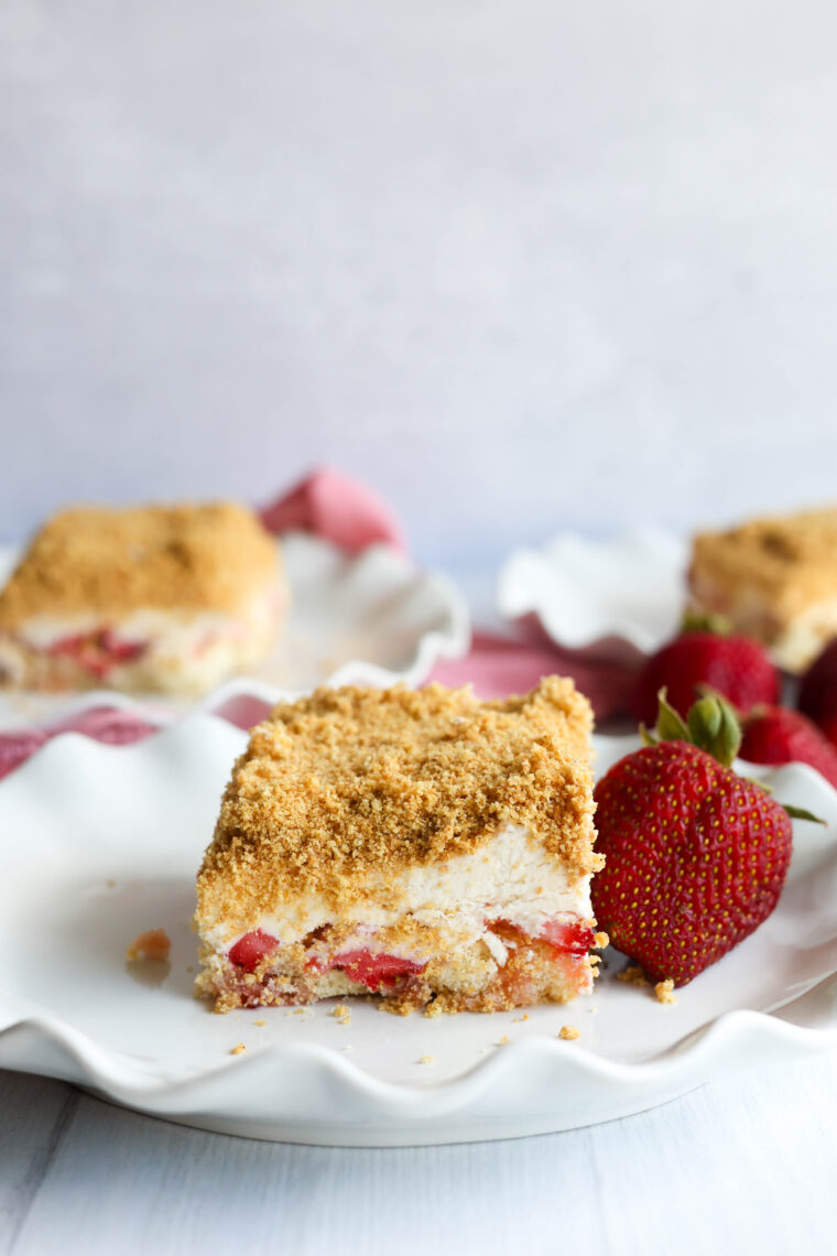 A slice of no-bake strawberry shortcake with crumbly topping sits on a white plate by a couple of fresh strawberries, while more tasty dessert bars hang out in the background.