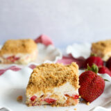 A slice of no-bake strawberry shortcake with crumbly topping sits on a white plate by a couple of fresh strawberries, while more tasty dessert bars hang out in the background.