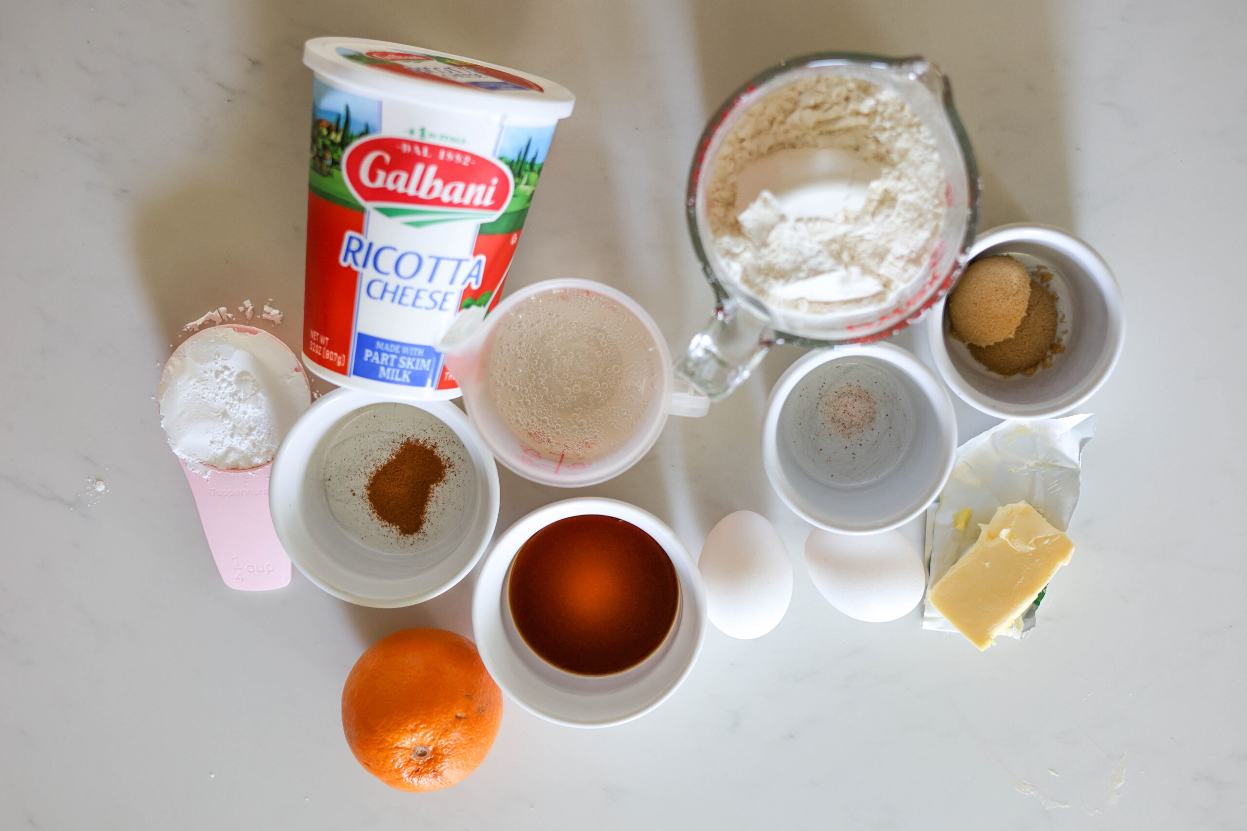 A bunch of baking basics—ricotta cheese, flour, white and brown sugar, eggs, butter, an orange, vanilla, and some spices—are spread out on a white countertop.