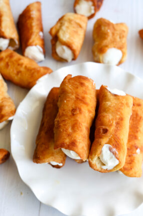 A plate piled with golden-brown cannoli filled with bright orange ricotta looks super tempting, while a few more cannoli are scattered around on a white surface in the background.
