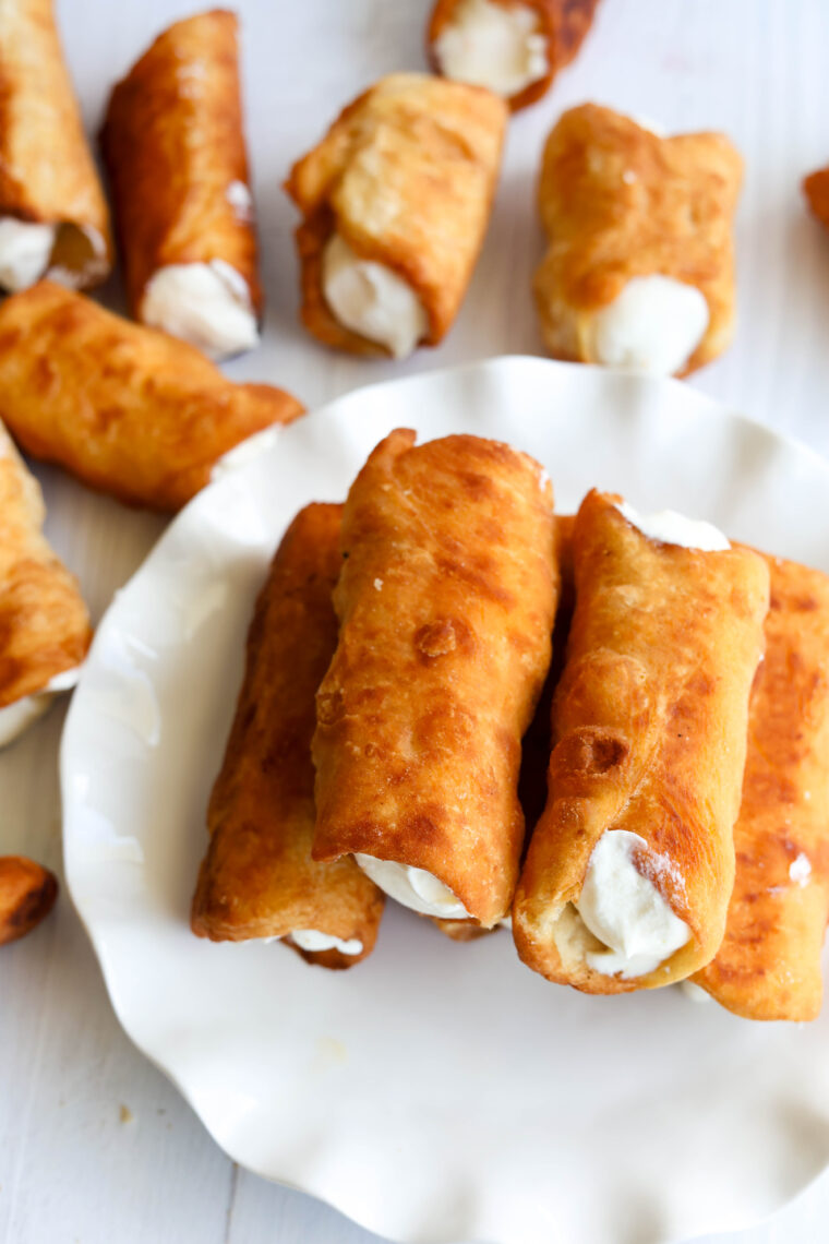 A plate piled with golden-brown cannoli filled with bright orange ricotta looks super tempting, while a few more cannoli are scattered around on a white surface in the background.