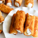 A plate piled with golden-brown cannoli filled with bright orange ricotta looks super tempting, while a few more cannoli are scattered around on a white surface in the background.