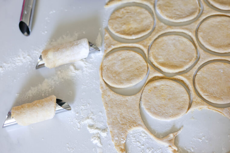 Circles of dough sit on a floured countertop, some wrapped around two metal pastry rollers. These are about to become tasty Cannoli cookies with orange ricotta inside or crispy treats after baking—yum!.
