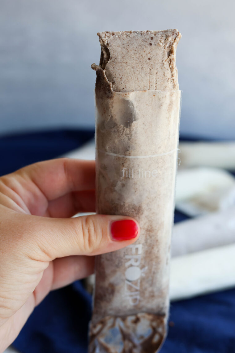 Someone with red nail polish is holding a half-eaten Ninja Slushi in a clear plastic tube, with the background all blurred out.