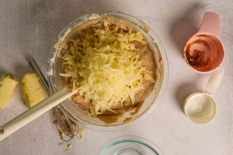A bowl of batter topped with grated apples, plus measuring cups, a whisk, and chopped apples on a light countertop.