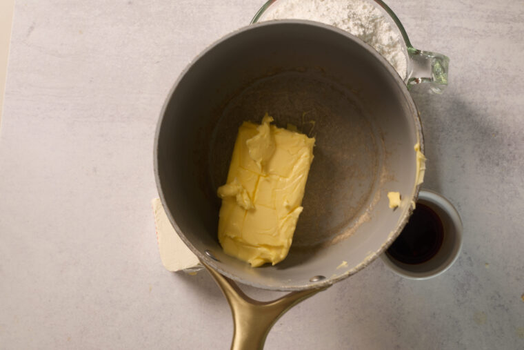 A pan with a stick of butter sits on a light counter, with a small cup of dark liquid—maybe vanilla—right beside it.