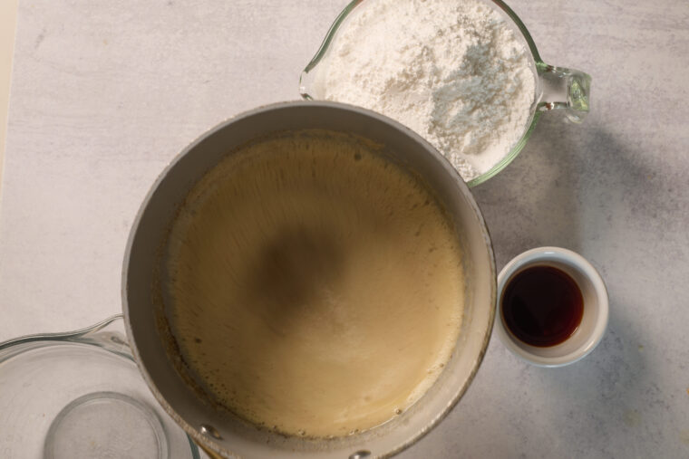 Mixing bowl with foamy batter, a dish of dark liquid, and flour on a light table—looks like cake-baking time!.