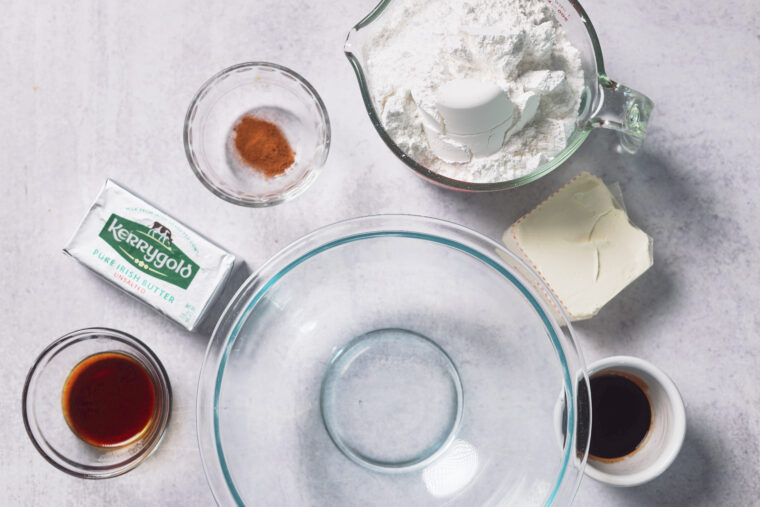 Overhead shot: flour with an egg, cream cheese, vanilla, cinnamon, butter, and an empty bowl on a light countertop.