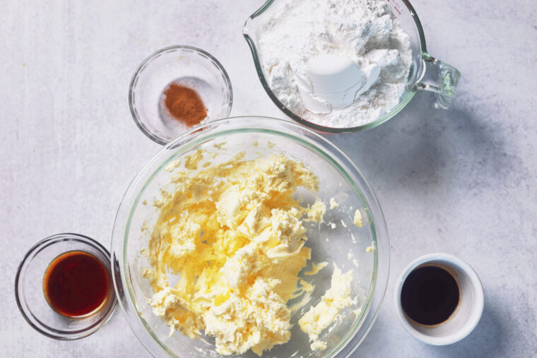 Creamed butter and sugar in a bowl, flour and egg in a cup, cinnamon, plus vanilla and extract bowls all set out on a light counter.