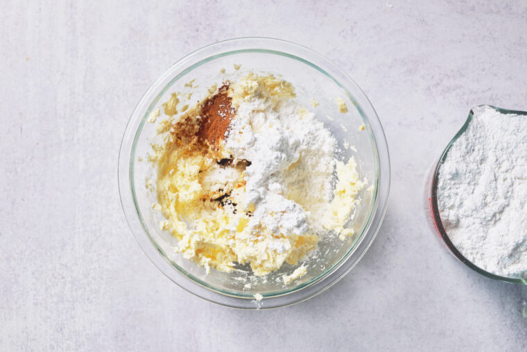 A glass bowl with creamed butter, sugar, flour, and vanilla sits on a light counter; a flour measuring cup peeks in above.
