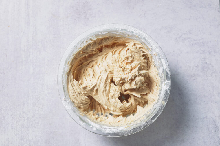 A glass bowl of fluffy light brown buttercream frosting sits on a gray surface, ready for your pumpkin cinnamon rolls.