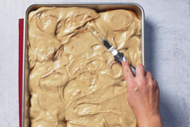 Someone’s spreading thick, pumpkin-cinnamon roll style frosting over a sheet cake with an offset spatula in a metal pan.
