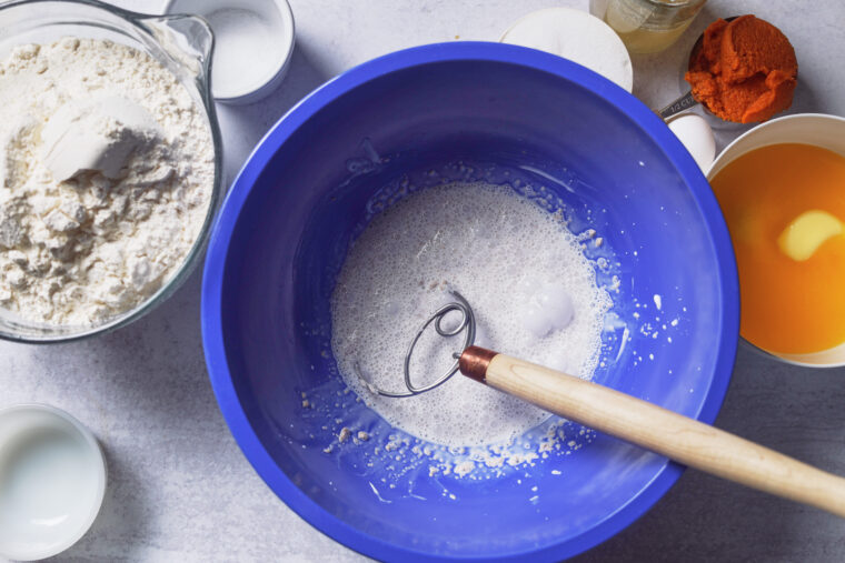 Blue bowl with whisk and bubbly mix, surrounded by bowls of flour, sugar, milk, pumpkin puree, and orange butter mix.
