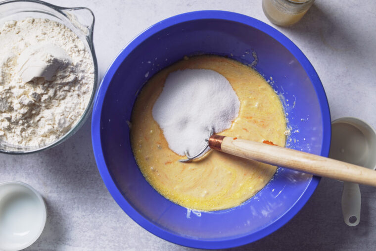 A blue bowl with yellow pumpkin batter getting stirred, sprinkled with sugar. Flour, milk bowls and a measuring cup sit close by.